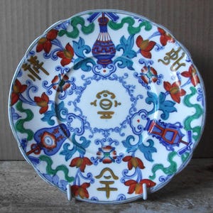 May include: A round ceramic plate featuring a white base with a colorful pattern of blue, green, and red floral motifs, stylized vases, and gold-colored Chinese characters. The design includes intricate blue scrollwork and symmetrical floral elements.