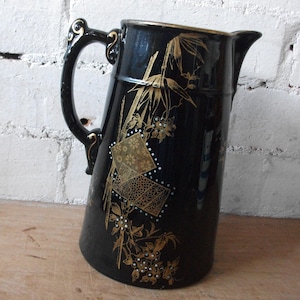 May include: Black ceramic pitcher with gold floral and bamboo design. The pitcher has a curved handle with gold accents and a spout for pouring. The design includes square and rectangular shapes with floral patterns.