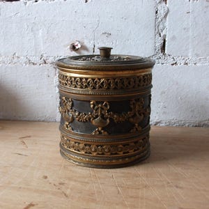 May include: A round, gold-coloured metal container with a decorative lid. The container has a black base with intricate gold designs.
