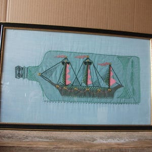 May include: A framed textile art piece featuring a light blue fabric background with an embroidered ship inside a bottle outline. The ship has dark green and pink sails, black rigging, and small gold-coloured circular accents along the hull.