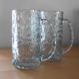 May include: Two clear glass beer mugs with textured sides and sturdy handles. The mugs are cylindrical with a thick base. The textured design gives the mugs a unique, rustic appearance. The mugs are empty and ready to be filled.