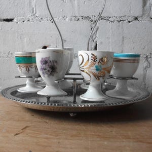 May include: A silver egg cup holder with six white porcelain egg cups. The egg cups have different floral and geometric designs in gold and turquoise.