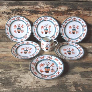 May include: A vintage porcelain dish set with a floral and vase motif. The set includes six plates and a small jug. The plates have a white base with a teal border and orange and teal details. The jug has a handle and a matching design.