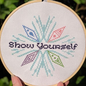 Pode incluir: Um bastidor de bordado em ponto cruz com as palavras "Show Yourself" em fio azul escuro. O desenho apresenta um padrão semelhante a um floco de neve com detalhes em azul, rosa, roxo e verde. O bordado está num bastidor de madeira.