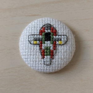 May include: A round, off-white fabric button featuring a cross-stitch design of a spaceship. The spaceship is primarily gray, with red, green, and black accents. Yellow details are visible on the wings. The button is set against a light wood grain background.