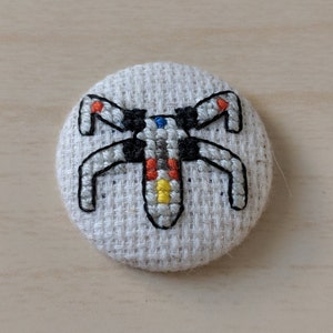 Op de afbeelding: Een wit, zwart, oranje, geel en blauw geborduurd ruimteschip op een beige stoffen knoop.