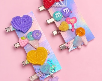 May include: A set of eight metal hair clips attached to two holographic display cards. The clips feature various colorful resin charms including a red strawberry, blue and purple letter m charms, hearts, a lollipop, and a light purple bear charm.