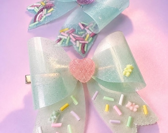 May include: Two hair bows made of translucent, glittery light blue material. The top bow features a purple circular center and colorful sprinkles. The bottom bow has a pink heart-shaped center, colorful sprinkles, and small bear-shaped charms.