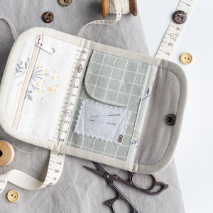 May include: An open, rectangular sewing kit with a zipper, pockets, and a snap closure. The interior features floral and grid patterns, with a ruler printed along the edge. A measuring tape, scissors, wooden spools, and buttons surround the kit.