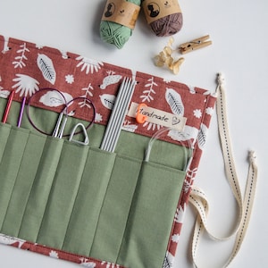 Puede incluir: Un estuche enrollable de tela verde y marrón con múltiples bolsillos para guardar agujas de tejer. El estuche tiene un patrón floral blanco y rojo y las palabras "hecho a mano" están impresas en una etiqueta. Dos ovillos de lana son visibles en el fondo.