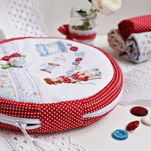 May include: A round, red and white polka dot fabric case with a zipper closure. The case is filled with white fabric and a cross-stitch design of a woman sewing. The case is sitting on a white surface with a white lace doily and red and blue buttons.