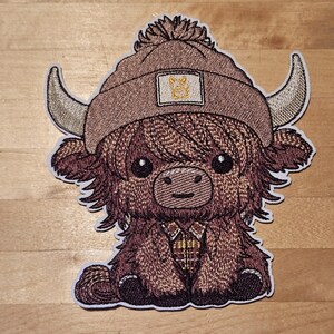 May include: Embroidered patch of a cartoon Highland cow wearing a brown beanie with a small patch. The cow has large eyes, a plaid shirt, and curved horns. The patch is outlined in white.