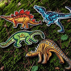 Peut inclure: Quatre écussons brodés de dinosaures comprenant un stégosaure jaune et orange, un théropode bleu, un sauropode vert à long cou et un dinosaure cuirassé beige avec des taches marron foncé, tous avec des bordures blanches et des coutures détaillées.
