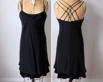 May include: A black slip dress with spaghetti straps and a layered hemline. The dress is displayed on a vintage dress form, showcasing the front and back details. The back features a unique crisscross strap design.