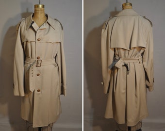 Op de afbeelding: Een beige trenchcoat op een paspop. De jas heeft een dubbele rij knopen, een ceintuur in de taille en knopen. Het heeft epauletten op de schouders en een split aan de achterkant. De stof is glad en mogelijk waterafstotend.