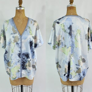 Può includere: Top azzurro a maniche corte con scollo a V e silhouette fluida. Il tessuto presenta un motivo ad acquerello nei toni del blu, grigio e verde. Il top è esposto su un manichino vintage.
