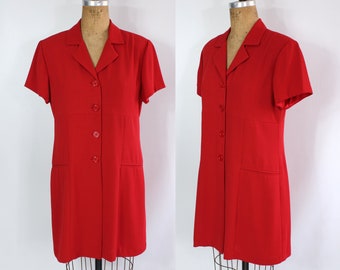 Op de afbeelding: Een felrode jurk met korte mouwen, een kraag en een knoopsluiting aan de voorkant. De jurk heeft twee voorzakken en wordt gepresenteerd op een vintage paspop. Een klassiek, op maat gemaakt kledingstuk.