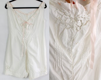 May include: White vintage romper with a V-neckline, adorned with pink ribbon and lace trim. The garment features delicate embroidery and pleating details, with a scalloped hemline. The romper is sleeveless and has a split front design.
