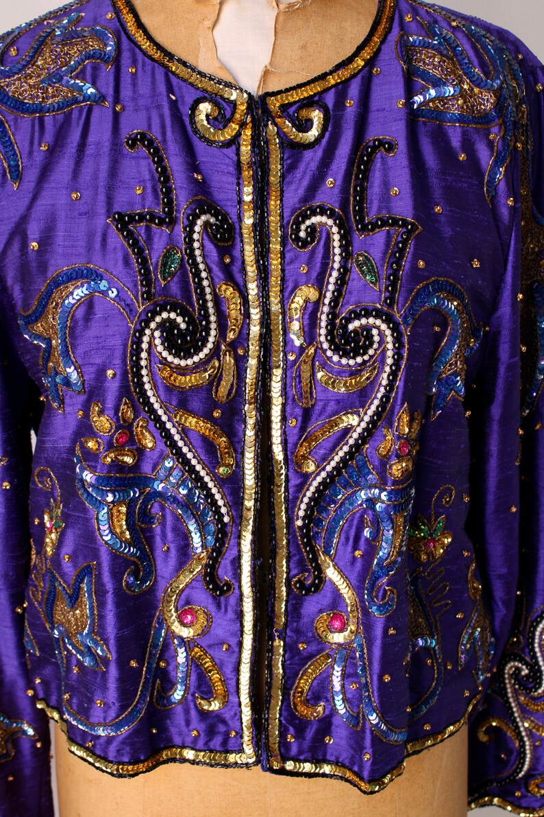 May include: A vibrant purple jacket with intricate embellishments. The jacket features gold sequins, blue and black beading, and decorative patterns. The design includes a round neckline and long sleeves. The jacket is a statement piece.
