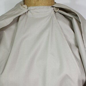 Op de afbeelding: Een lichtbeige cape met een ronde kraag en twee knoopsluitingen. De cape wordt getoond op een mannequin. De stof lijkt een lichtgewicht materiaal te zijn, geschikt voor layering. Het ontwerp van de cape is eenvoudig en klassiek.