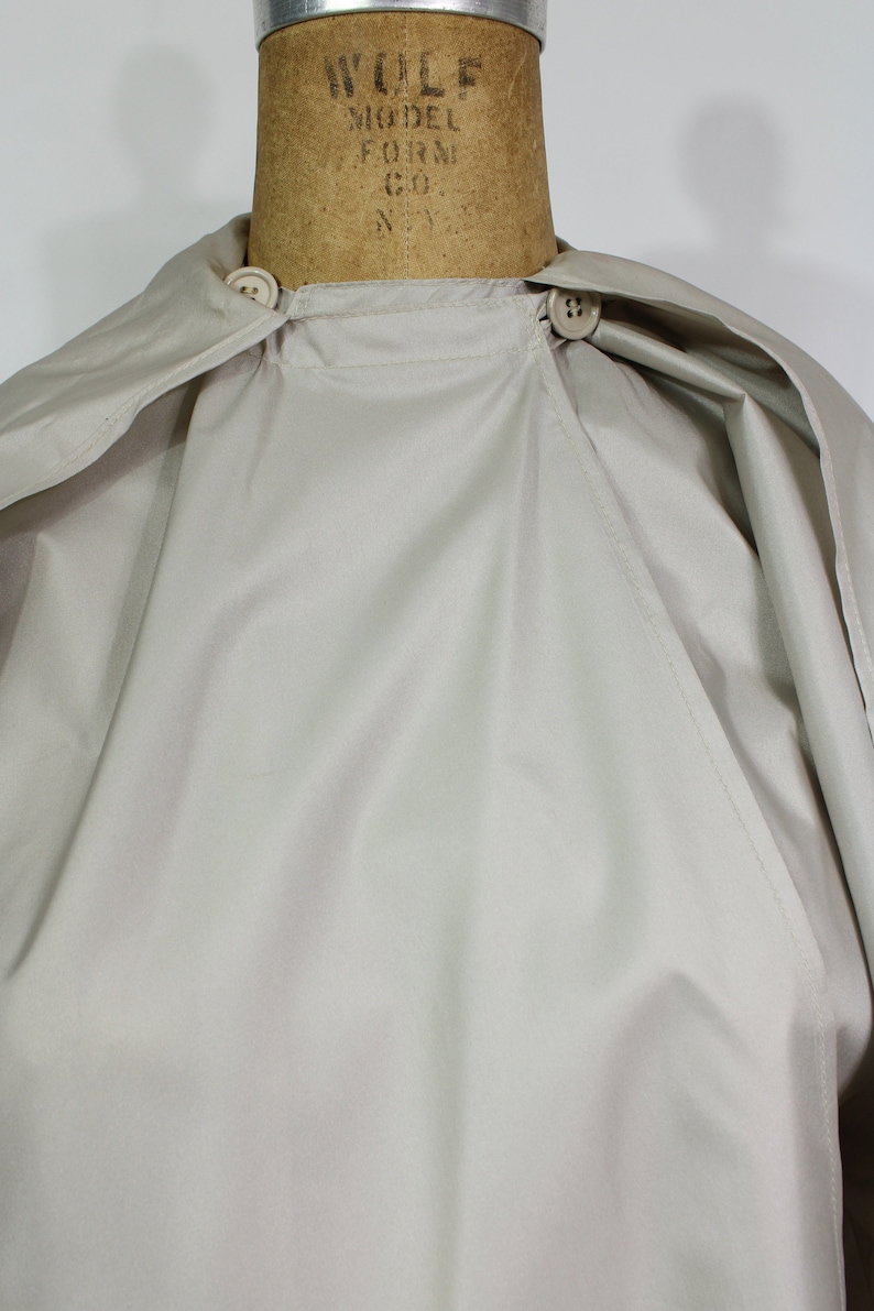 Op de afbeelding: Een lichtbeige cape met een ronde kraag en twee knoopsluitingen. De cape wordt getoond op een mannequin. De stof lijkt een lichtgewicht materiaal te zijn, geschikt voor layering. Het ontwerp van de cape is eenvoudig en klassiek.
