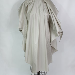 Op de afbeelding: Een lichtbeige cape-achtig kledingstuk, gedrapeerd over een vintage paspop. De cape heeft een brede, afgeronde kraag en valt in zachte plooien. De paspop staat op een zwarte metalen standaard met wielen, tegen een witte achtergrond.