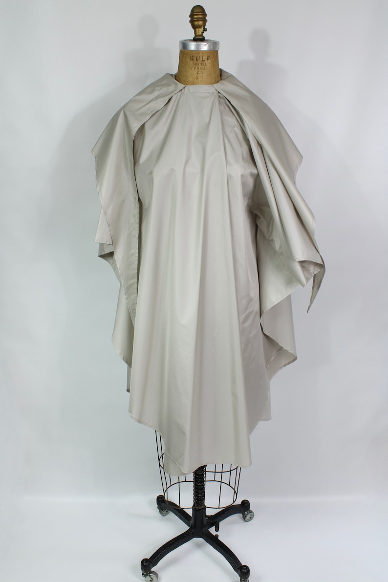 Op de afbeelding: Een lichtbeige cape-achtig kledingstuk, gedrapeerd over een vintage paspop. De cape heeft een brede, afgeronde kraag en valt in zachte plooien. De paspop staat op een zwarte metalen standaard met wielen, tegen een witte achtergrond.