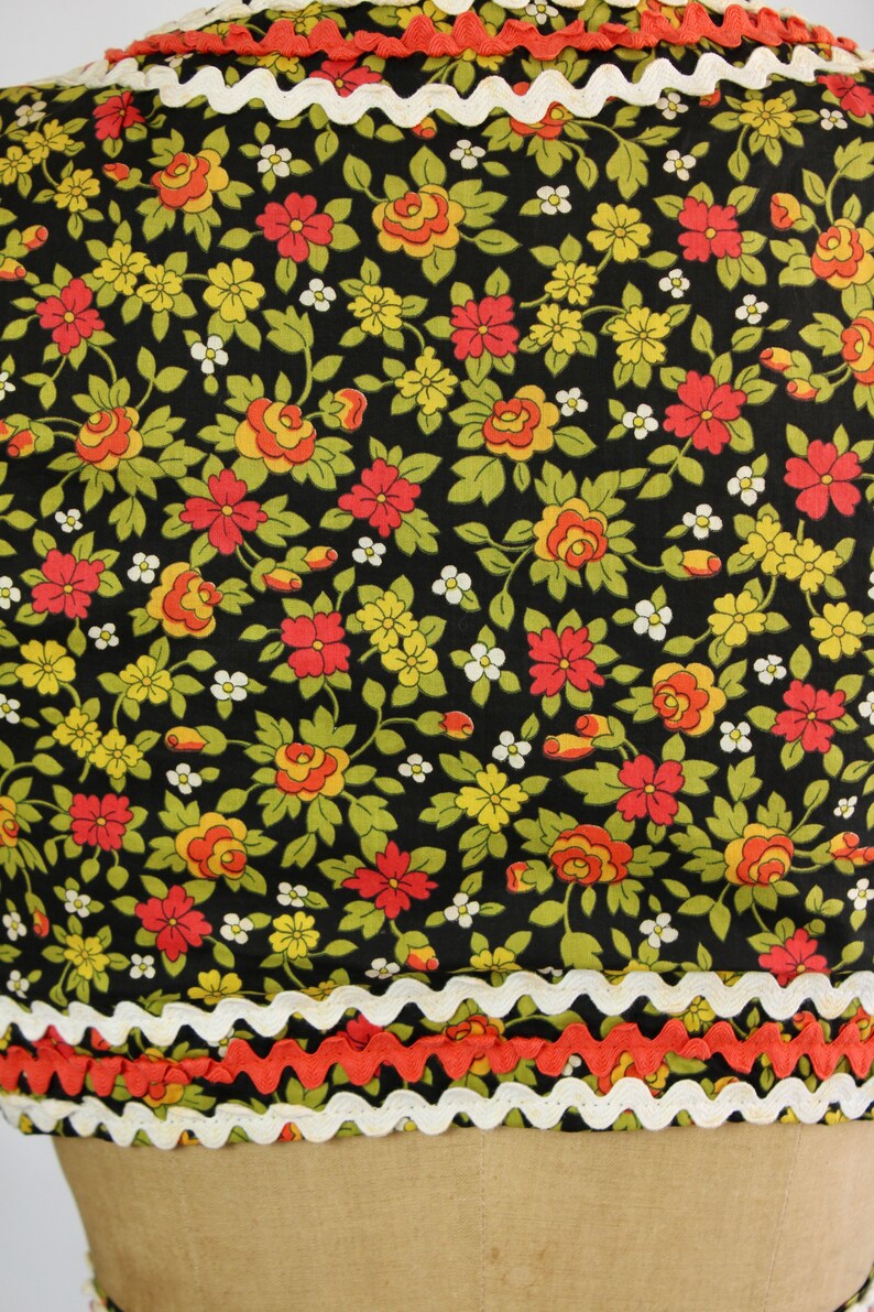 May include: Close-up of a floral fabric with a black background. The fabric features red, yellow, and white flowers with green leaves. The edges are trimmed with white and orange zigzag trim, creating a decorative border.