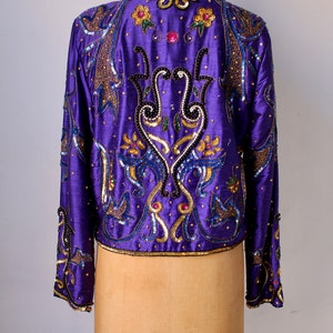 May include: A vibrant purple jacket with intricate gold, blue, and black sequin and bead embellishments. The jacket features floral and abstract patterns, with long sleeves and a round neckline. The garment is displayed on a mannequin.