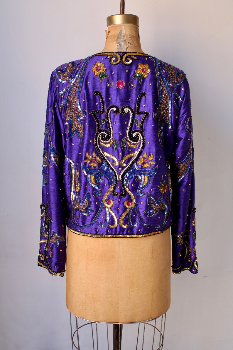 May include: A vibrant purple jacket with intricate gold, blue, and black sequin and bead embellishments. The jacket features floral and abstract patterns, with long sleeves and a round neckline. The garment is displayed on a mannequin.