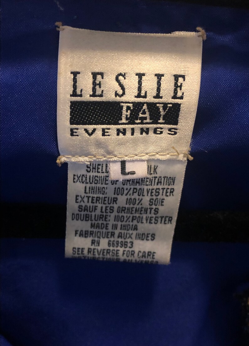 May include: A white tag with the text "LESLIE FAY EVENINGS" in black. The tag also includes fabric content details, size "L", and "MADE IN INDIA". The tag is against a blue fabric.