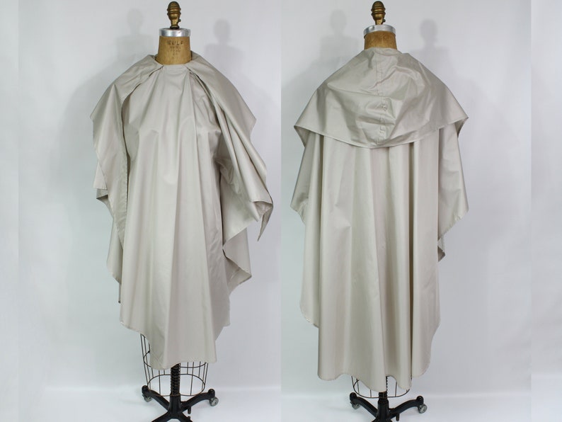 Op de afbeelding: Een lichtbeige saloncape, getoond op een vintage paspop. De cape heeft een breed, gedrapeerd ontwerp met een kraag en een capuchon. De stof lijkt een glad, lichtgewicht materiaal te zijn, geschikt voor professioneel gebruik in een kapsalon of spa.
