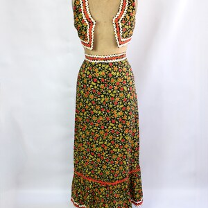 May include: A vintage two-piece outfit featuring a vest and a maxi skirt. The black fabric is covered in a floral pattern with red, yellow, and green flowers. The vest and skirt have orange and white trim. The outfit is displayed on a mannequin.