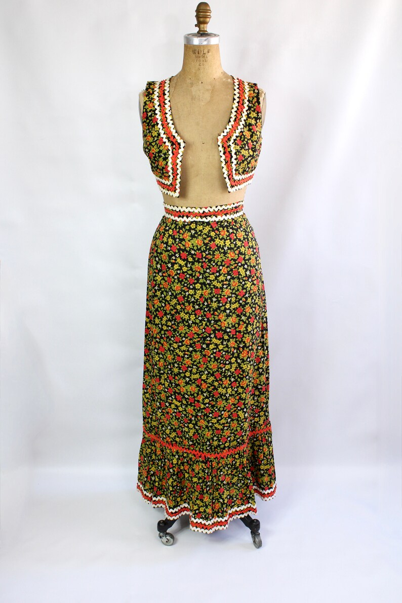 May include: A vintage two-piece outfit featuring a vest and a maxi skirt. The black fabric is covered in a floral pattern with red, yellow, and green flowers. The vest and skirt have orange and white trim. The outfit is displayed on a mannequin.