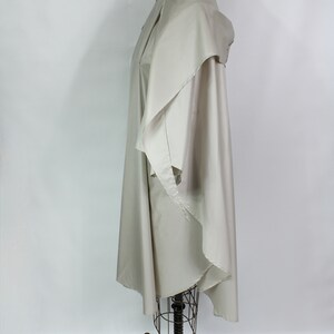 Op de afbeelding: Een lichtbeige cape-achtig kledingstuk met een gedrapeerde kraag en een vloeiend ontwerp. De cape wordt getoond op een vintage paspop met een zwarte metalen basis en wielen. De stof lijkt licht van gewicht.