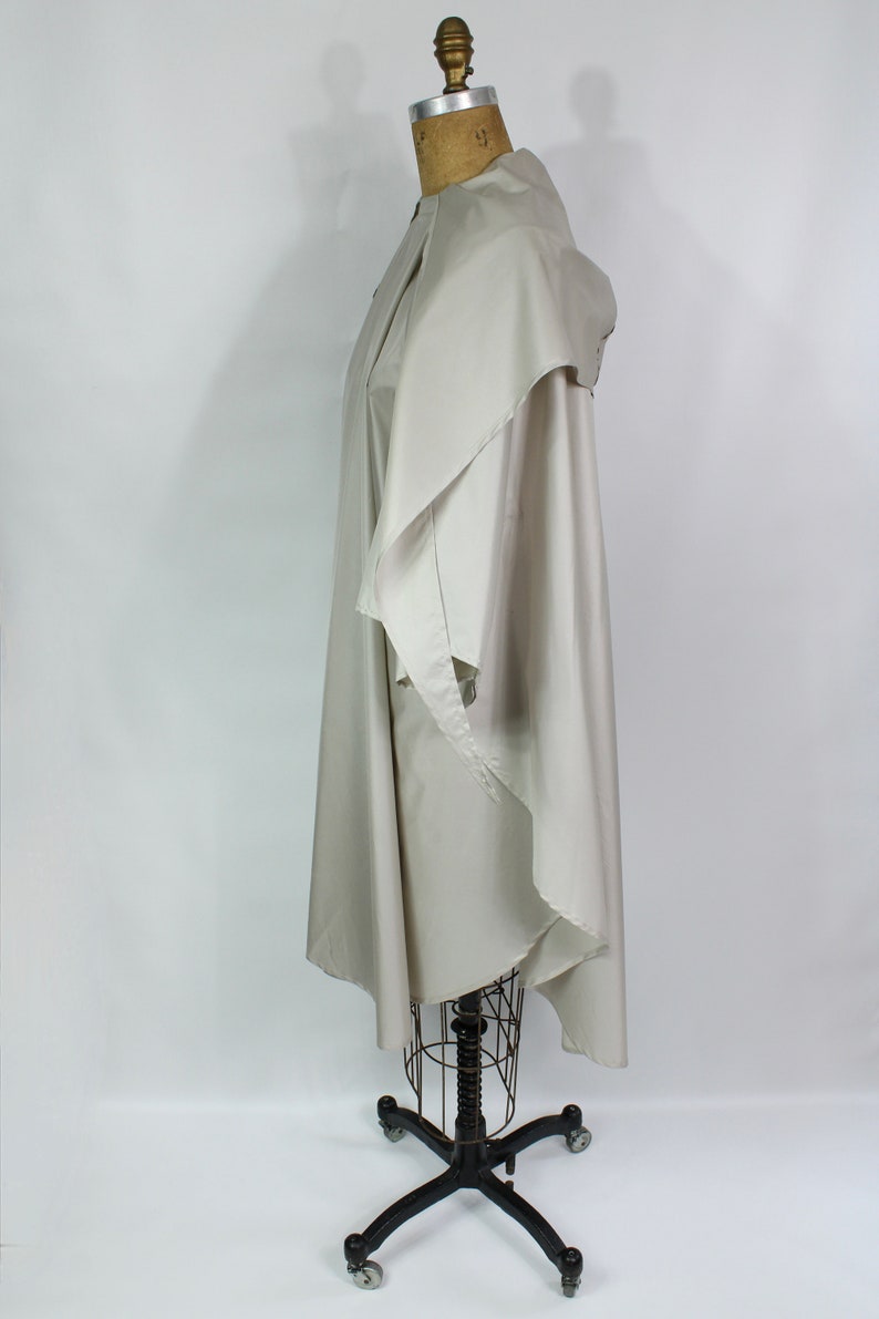 Op de afbeelding: Een lichtbeige cape-achtig kledingstuk met een gedrapeerde kraag en een vloeiend ontwerp. De cape wordt getoond op een vintage paspop met een zwarte metalen basis en wielen. De stof lijkt licht van gewicht.