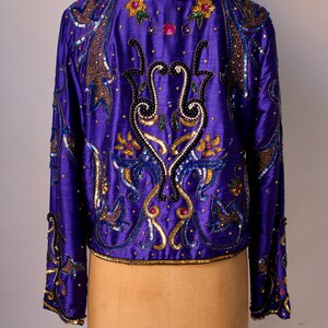 May include: A vibrant purple jacket with intricate sequin and bead embellishments. The jacket features gold, blue, and pink accents in a floral and abstract design. The long-sleeved jacket is displayed on a mannequin.