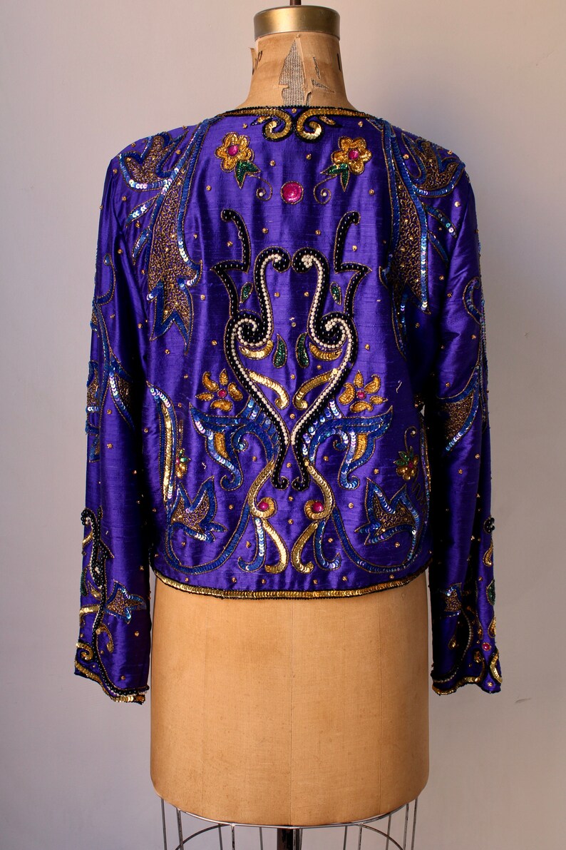 May include: A vibrant purple jacket with intricate sequin and bead embellishments. The jacket features gold, blue, and pink accents in a floral and abstract design. The long-sleeved jacket is displayed on a mannequin.