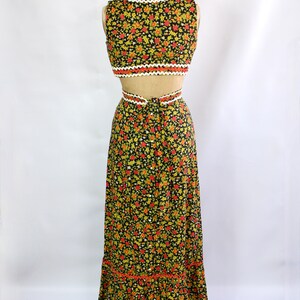 May include: A two-piece floral print outfit on a mannequin. The black fabric is covered in small red, yellow, and green flowers. The top is a crop top with a scalloped white and orange trim. The skirt is a maxi skirt with a matching trim.