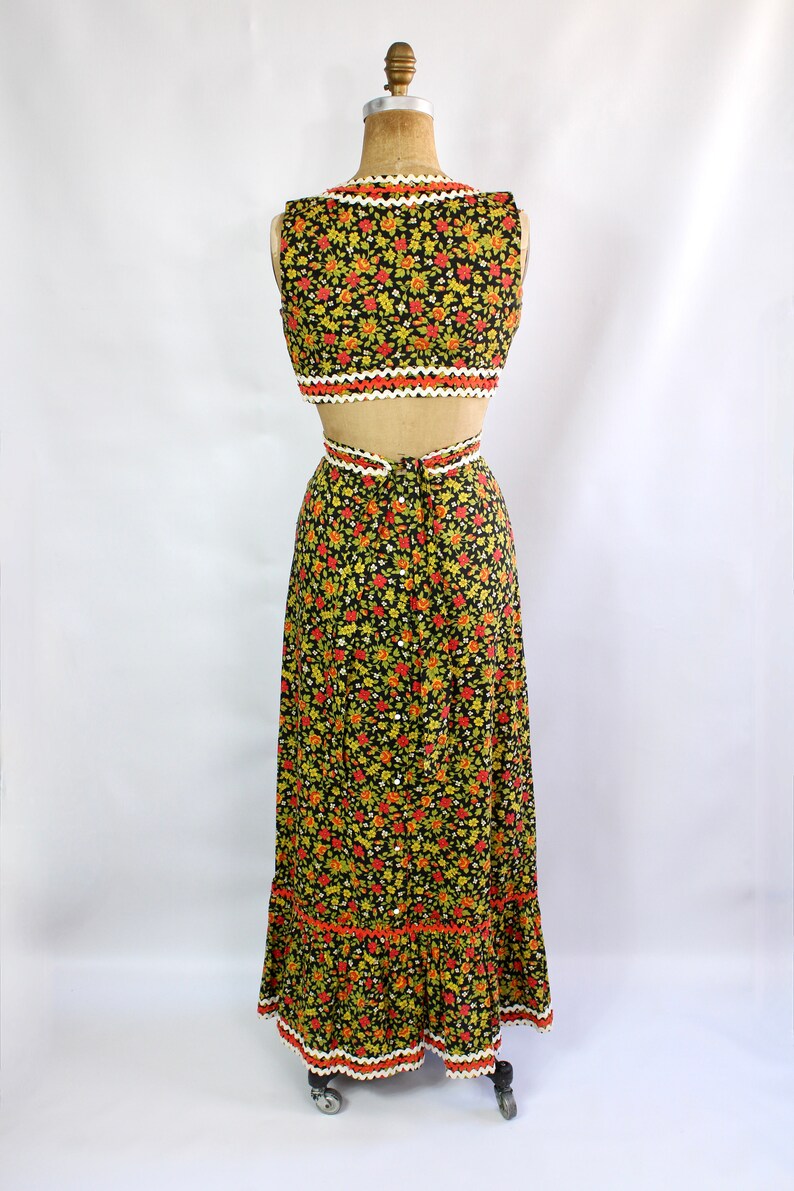 May include: A two-piece floral print outfit on a mannequin. The black fabric is covered in small red, yellow, and green flowers. The top is a crop top with a scalloped white and orange trim. The skirt is a maxi skirt with a matching trim.