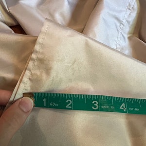 Op de afbeelding: Een close-up van een beige stof die wordt gemeten met een groen meetlint. De tape toont een meting van ongeveer 7,6 cm. De stof lijkt een licht, mogelijk satijnachtig materiaal te zijn.