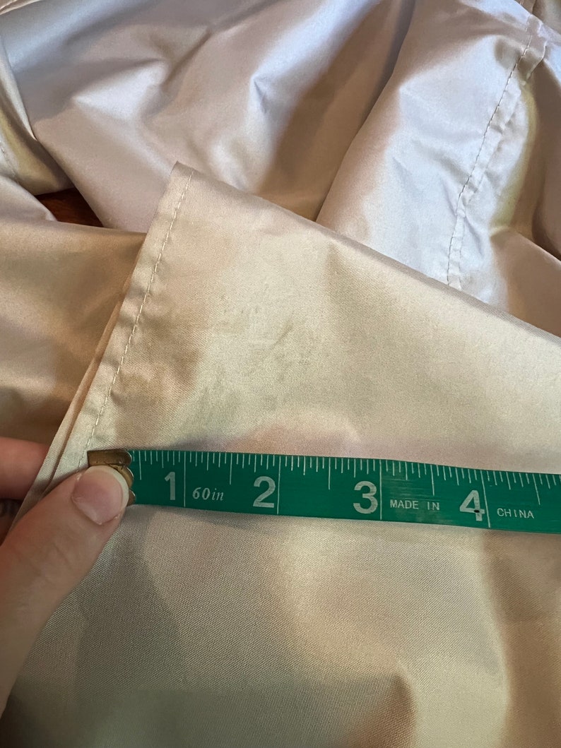 Op de afbeelding: Een close-up van een beige stof die wordt gemeten met een groen meetlint. De tape toont een meting van ongeveer 7,6 cm. De stof lijkt een licht, mogelijk satijnachtig materiaal te zijn.