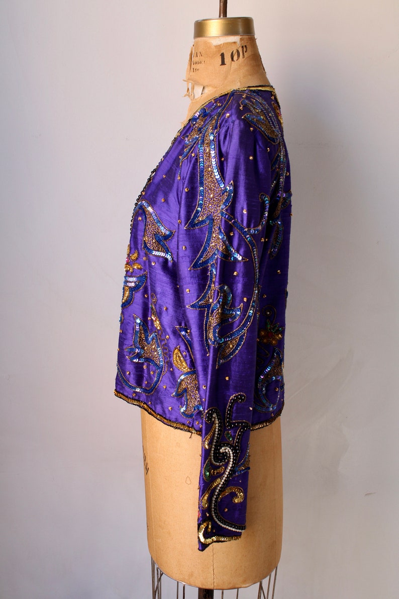 May include: A vintage purple jacket with intricate sequin and bead embellishments. The jacket features a detailed floral design in gold and blue, with a satin-like texture. The sleeves and collar are also adorned with the same decorative elements.