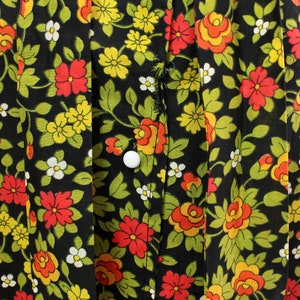 May include: Close-up of a black fabric with a floral pattern. The design features red, yellow, and orange flowers with green leaves and white accents. White buttons are visible on the fabric.