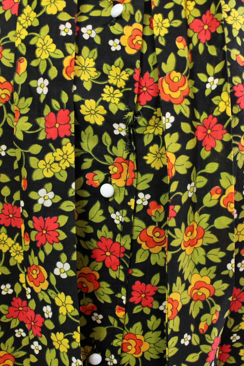 May include: Close-up of a black fabric with a floral pattern. The design features red, yellow, and orange flowers with green leaves and white accents. White buttons are visible on the fabric.