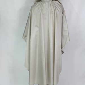 Op de afbeelding: Een lichtbeige poncho met capuchon, weergegeven op een zwarte metalen paspop. De poncho heeft een los, vloeiend ontwerp met een capuchon en een afgeronde zoom. De stof lijkt een lichtgewicht, waterbestendig materiaal te zijn, geschikt voor bovenkleding.