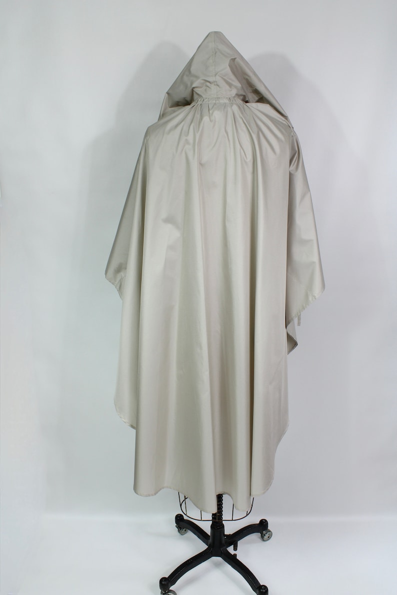 Op de afbeelding: Een lichtbeige poncho met capuchon, weergegeven op een zwarte metalen paspop. De poncho heeft een los, vloeiend ontwerp met een capuchon en een afgeronde zoom. De stof lijkt een lichtgewicht, waterbestendig materiaal te zijn, geschikt voor bovenkleding.