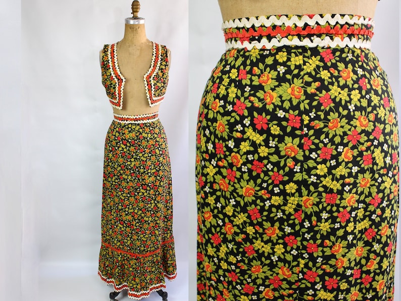 May include: Vintage two-piece floral set featuring a vest and a long skirt. The black fabric is covered in a pattern of red, yellow, and green flowers. The edges of the vest and skirt are trimmed with white and orange crochet detailing. The skirt measures approximately 40 inches in length.
