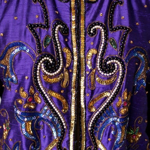 May include: Close-up of a purple jacket with intricate embellishments. The jacket features gold sequins, black beading, and blue and gold embroidered details. The design includes floral and swirling patterns, creating a luxurious and ornate appearance.