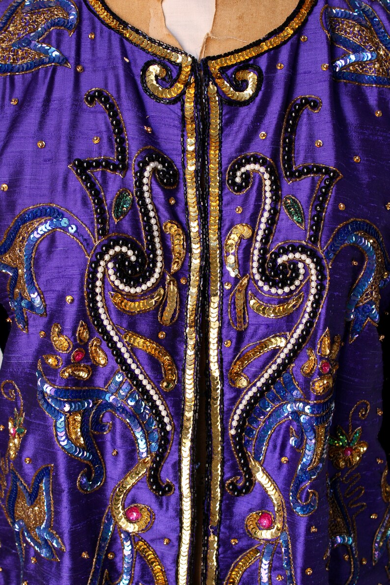 May include: Close-up of a purple jacket with intricate embellishments. The jacket features gold sequins, black beading, and blue and gold embroidered details. The design includes floral and swirling patterns, creating a luxurious and ornate appearance.